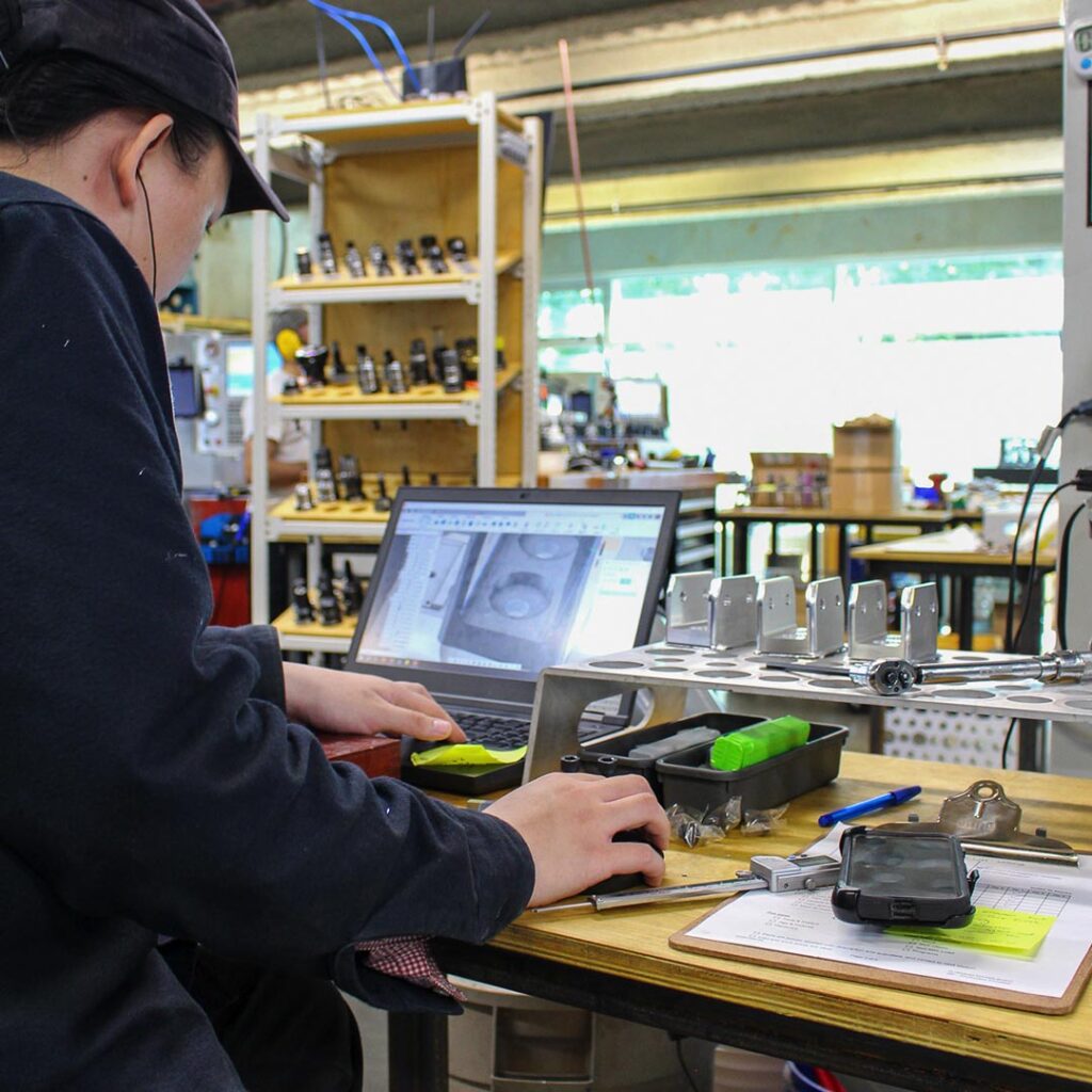 Focus Manufacturing shop floor 02 Photograph of a computer user on the shop floor. Digital calipers, a torque wrench, tool racks, a laptop, tool cases and a clipboard are visible. Background is brightly lit by sunlight.